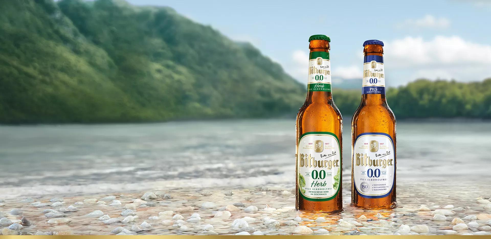 zwei Bitburger 0,0 Flaschen stehen am Ufer im Wasser vor einer bergigen Kulisse