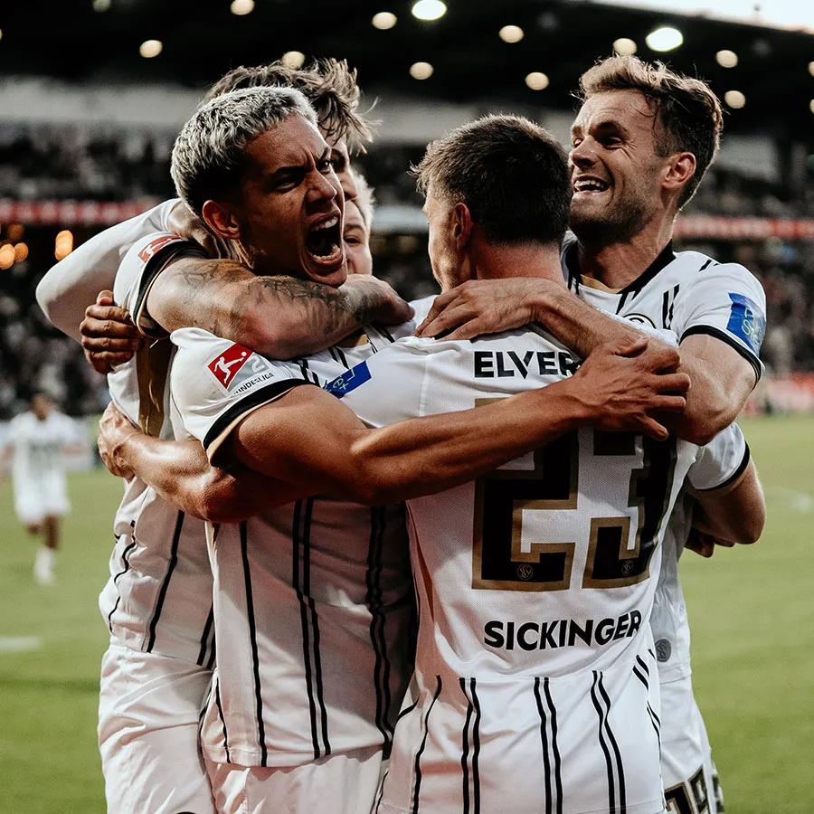Vier Spieler SV 07 Elversberg stehen auf einem Spielfeld und umarmen sich.