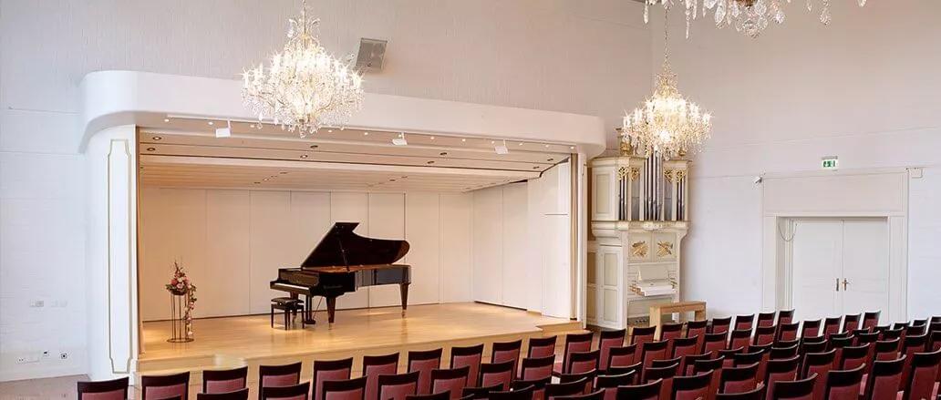 Scharzer Klavierflügel auf einer Bühne in einem weißen Raum, davor rote Stuhlreihen, der Saal verziert mit Stuck und Kronleuchter