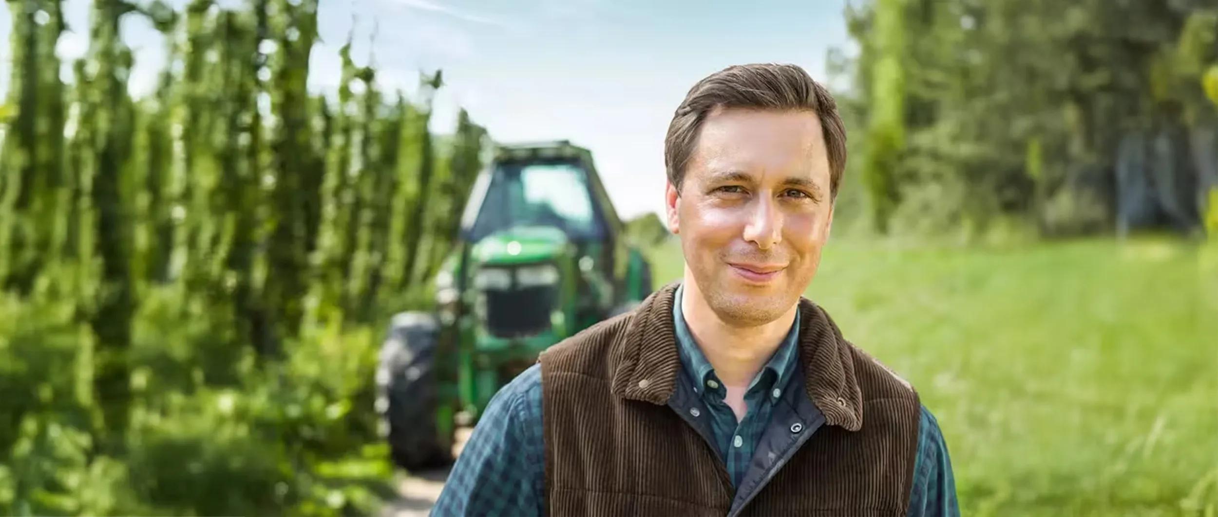 Eine Person steht im Freien vor einem grünen Traktor und üppigem Grün.