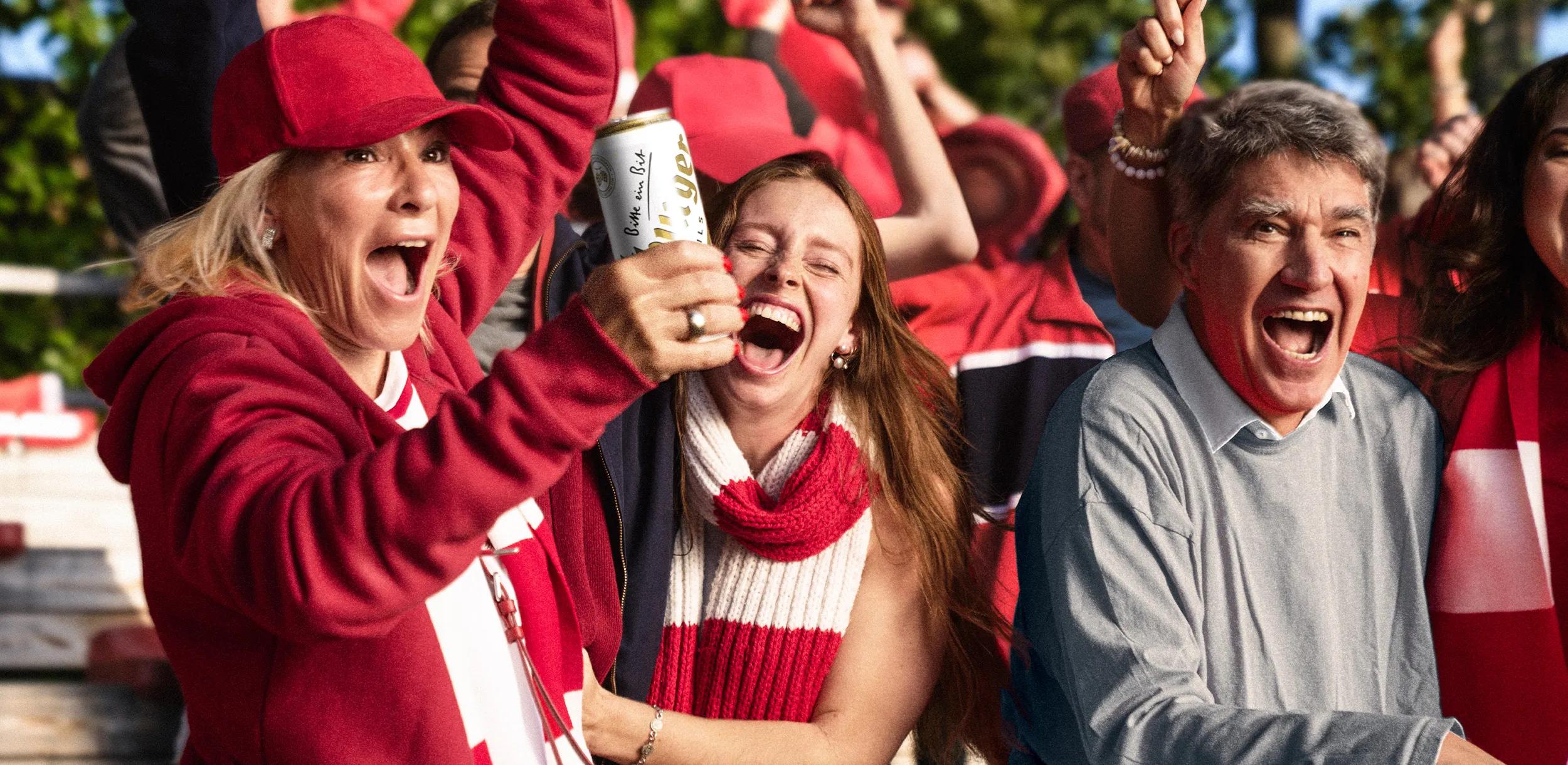 Drei Personen jubeln bei einem Fußballspiel, eine davon hält ein Bier in die Höhe.