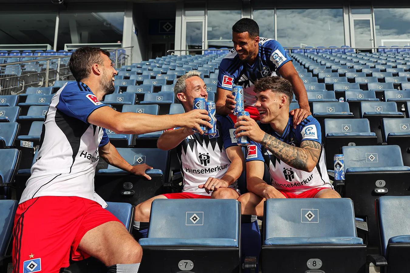 Vier Fußballspieler sitzen auf der leeren Tribüne und stoßen mit einem König Pilsener an.