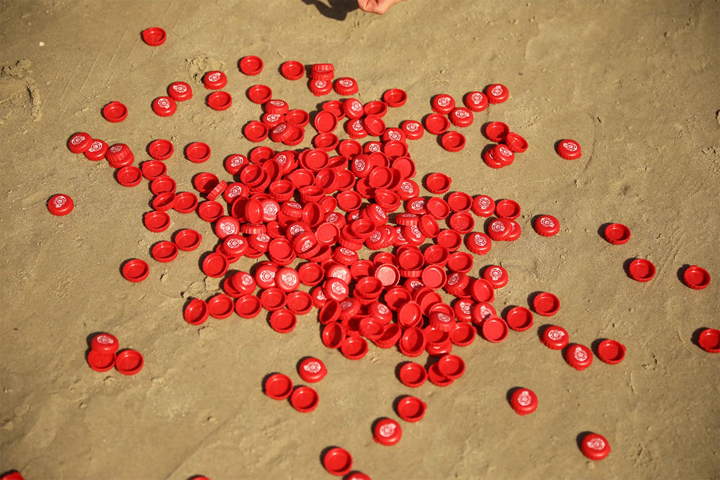 Rote Kronkorken mit Logo im Sand verstreut.