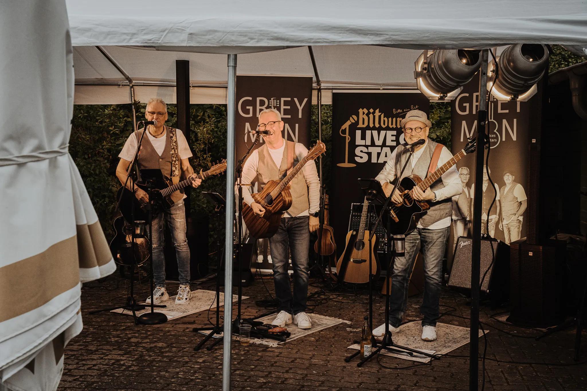 Drei Personen in Weste und Gitarre singen unter einem Partyzelt.