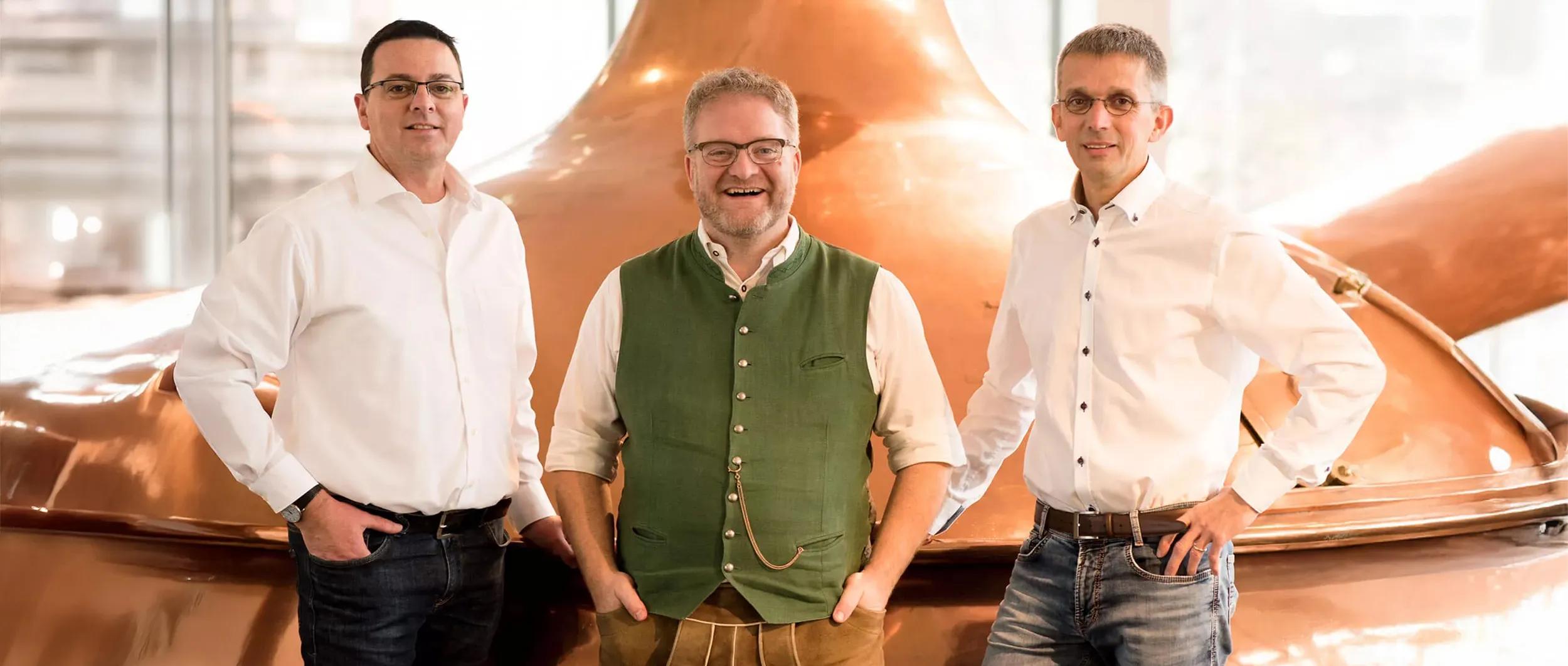 Drei Braumeister stehen in einem Sudhaus vor glänzenden Kupferkesseln.
