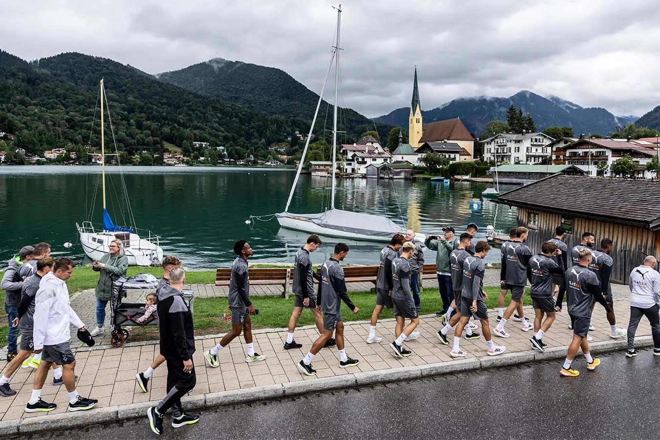 Foto vom Trainingslager Borussia Mönchengladbach. Spieler laufen auf einem Gehweg neben einem See entlang. Im Hintergrund ist eine bergige Landschaft zu sehen.