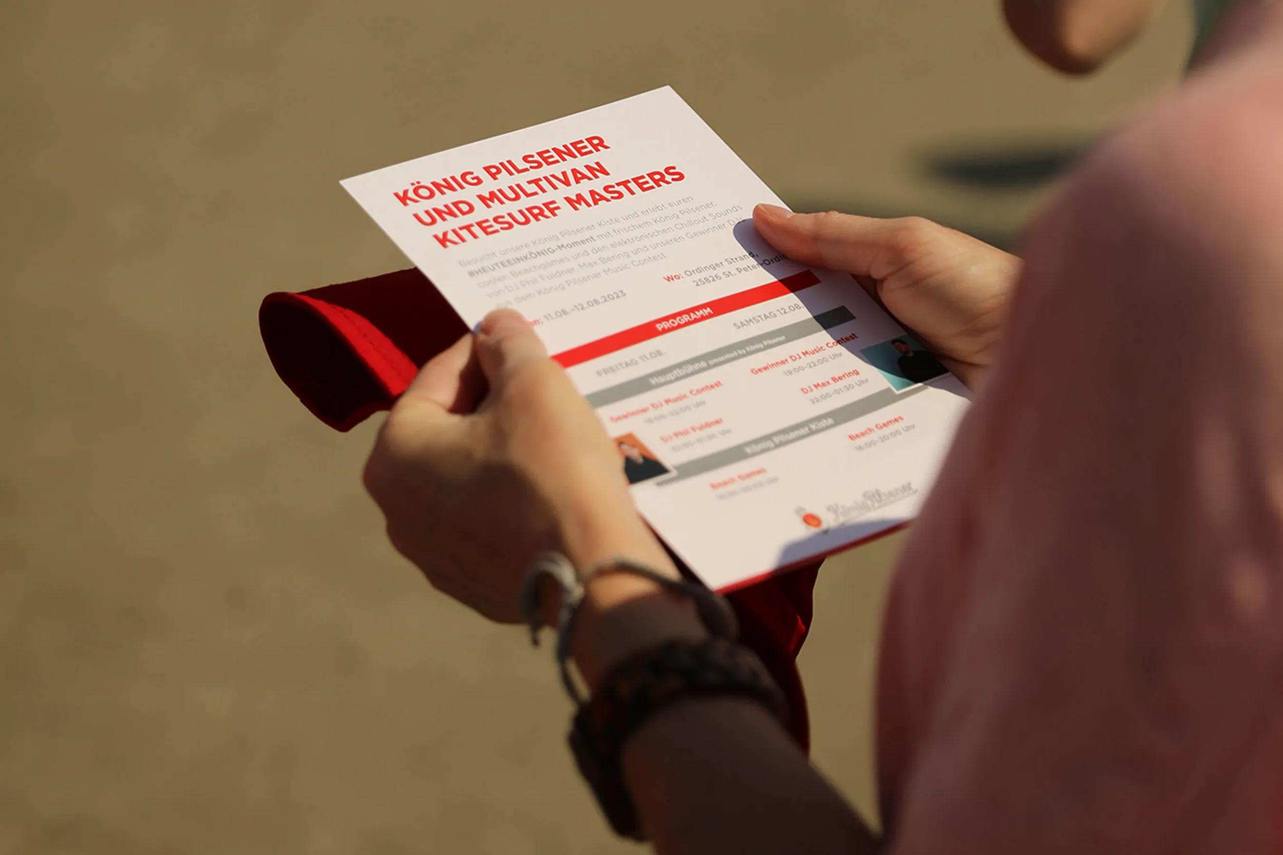 Nahaufnahme einer Hand, die ein Infoblatt zum „König Pilsener und Multivan Kitesurf Masters“-Event hält, auf dem das Programm für das Wochenende zu sehen ist.