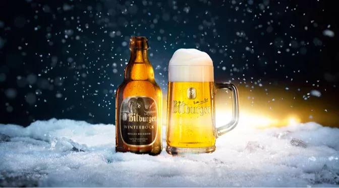 Stubbi Flasche Winterbock mit einem Bierkrug gefüllt mit Bitburger Winterbock auf schneebedecktem Boden, im Hintergrund Nachthimmel und es schneit.