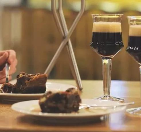 Zwei Gläser mit dunklem Bier und cremiger Schaumkrone stehen auf einem Holztisch. Daneben befinden sich Teller mit einem Stück Schokoladenkuchen. Eine Person schneidet mit einer Gabel ein Stück ab. Die Szene wirkt gemütlich und genussvoll.
