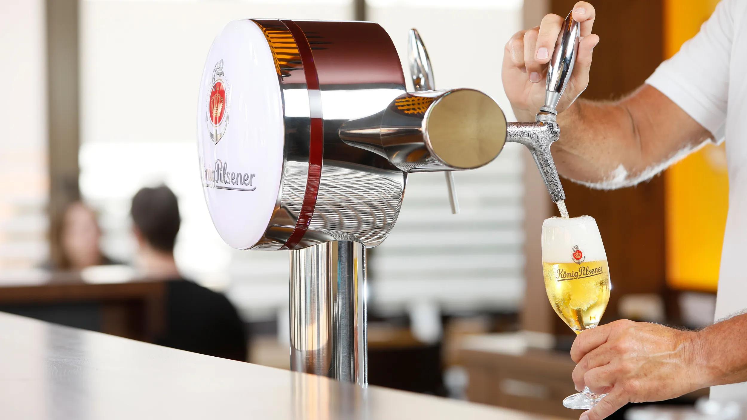 Im Vordergrund sieht man einen silbernen Zapfhahn mit König Pilsener Branding hinter einer Theke. Eine Person zapft ein Bier in eine Bier-Tulpe. Der Hintergrund ist verschwommen und man sieht unscharf zwei Personen an einem Tisch sich gegenübersitzend unterhalten. 