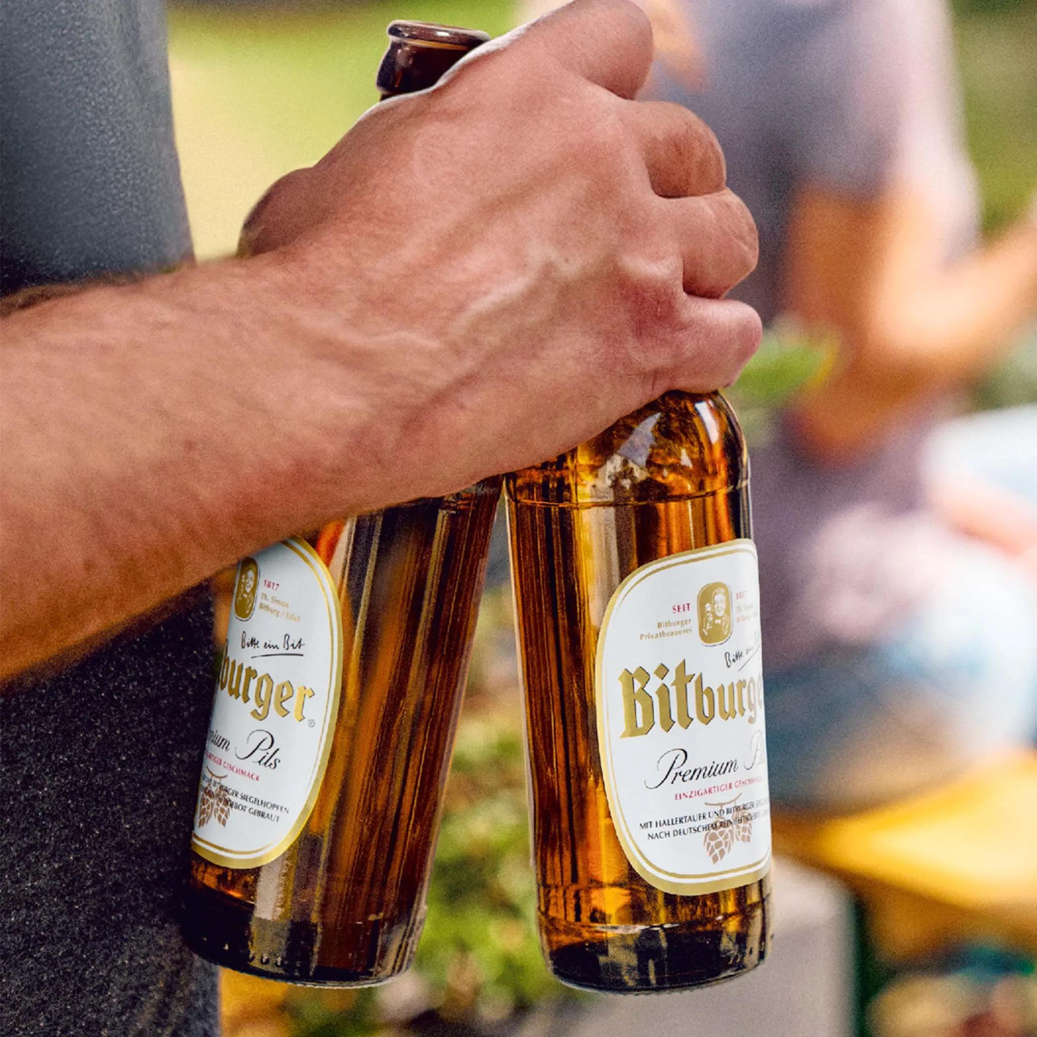 Eine Hand hält zwei Flaschen Bitburger Premium Pils, im unscharfen Hintergrund sitzen Menschen an einer Biergartengarnitur.