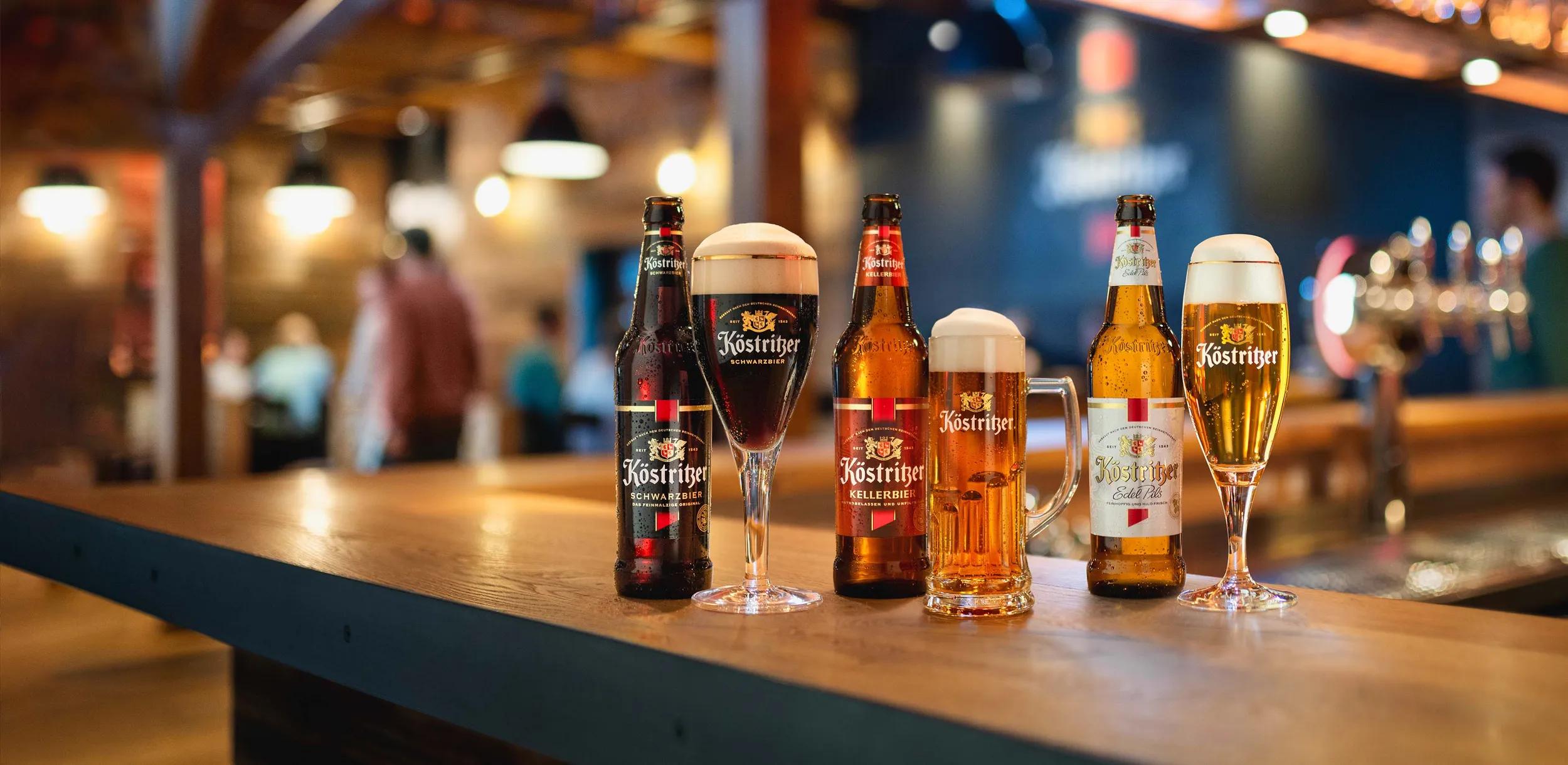 Verschiedene Köstritzer Biersorten in passenden Gläsern auf einer Holztheke in gemütlicher Bar-Atmosphäre.