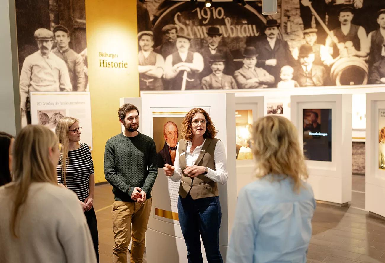 Eine Besuchergruppe steht im Raum in der Bitburger Erlebniswelt. Im Hintergrund ist eine Bildergalerie zu der Bitburger Geschichte zu sehen. An der Wand ist ein Schriftzug "Bitburger Historie".