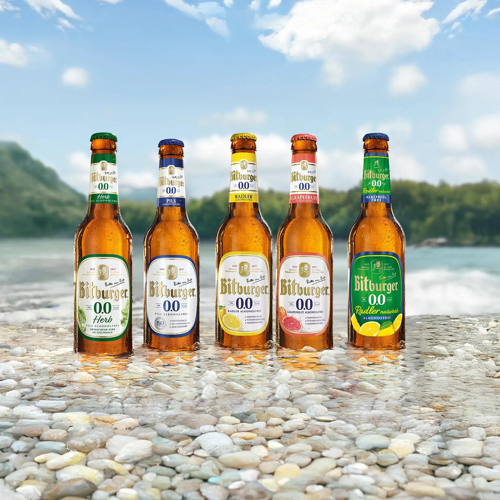 Fünf Sorten Bitburger alkoholfrei stehen nebeneinander im Wasser auf einem Kiesbett am See. Im Hintergrund ist eine hügelige Waldlandschaft zu sehen.