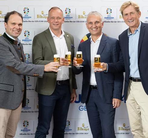 Pressemitteilung 4 Männer im Anzug gekleidet halten ein mit Bier gefülltes Pilsglas  vor der offiziellen EM Logo Wand 
