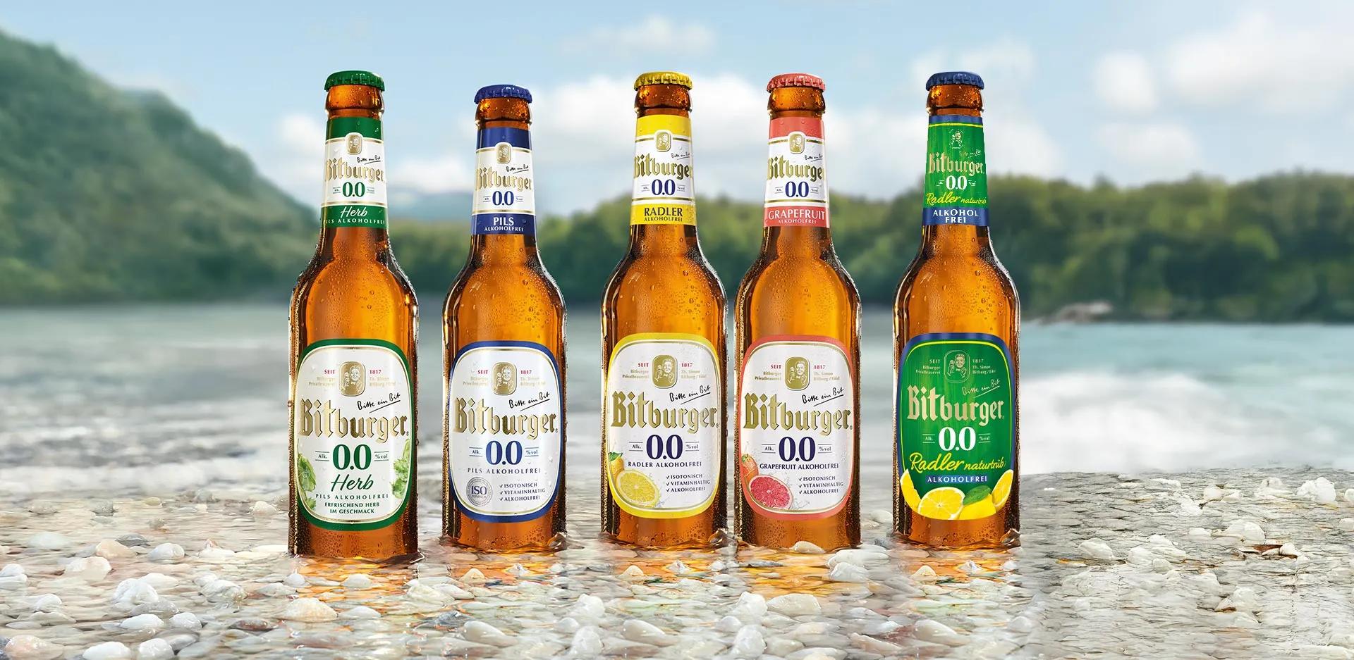 Eine Auswahl der Range von alkoholfreien Bitburger Bieren: Bitburger 0,0% Herb, Bitburger 0,0% Pils, Bitburger 0,0% Radler alkoholfrei, Bitburger 0,0% Grapefruit, Bitburger 0,0% Radler Naturtrüb. Die Flaschen stehen auf einem steinigen Untergrund am Strand, dezent im Wasser. Im Hintergrund sieht man einen hügeligen Waldrand vor einem See.