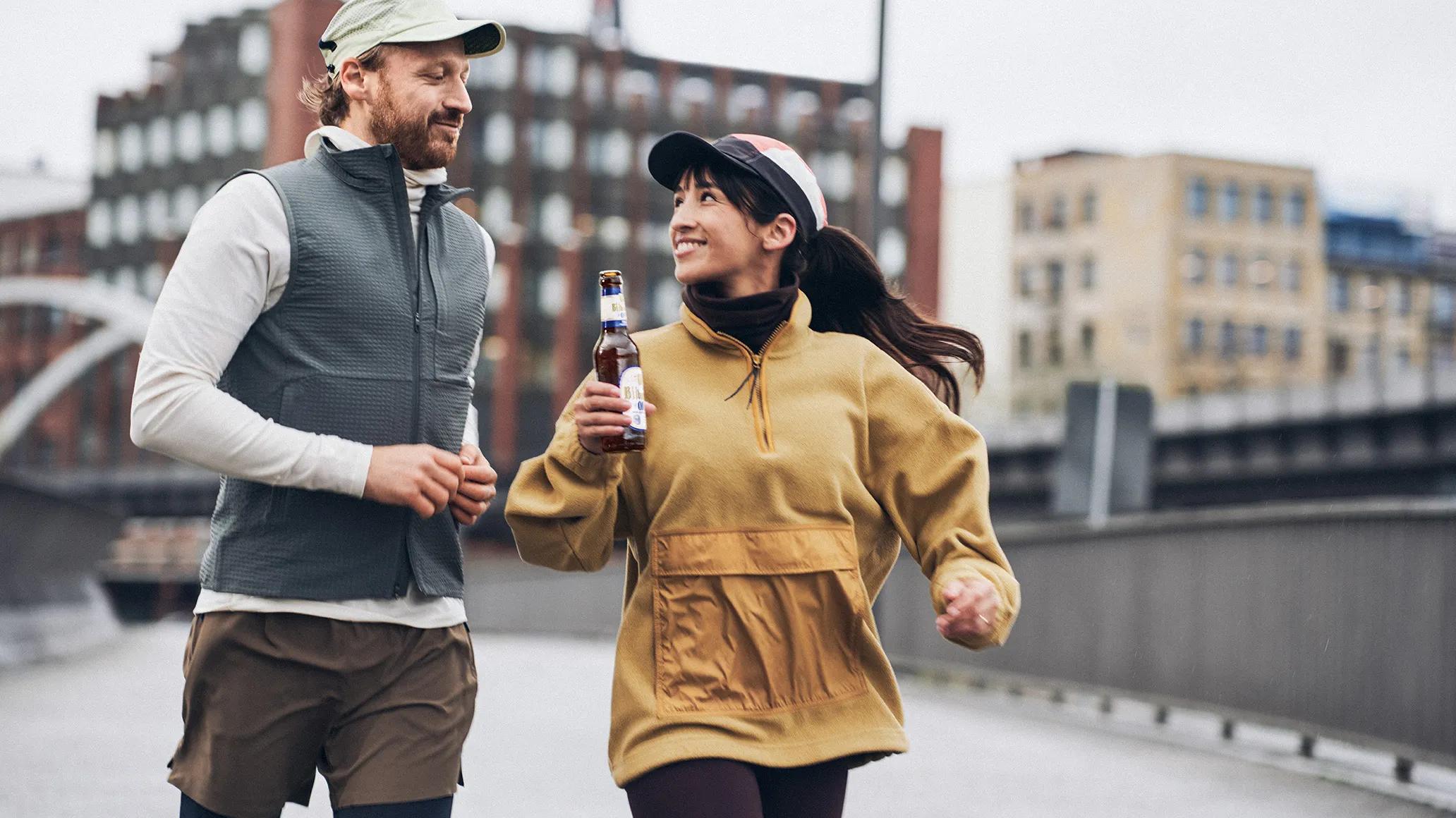 Zwei Läufer_innen gehen nebeneinander und trinken dabei ein Bitburger 0,0%