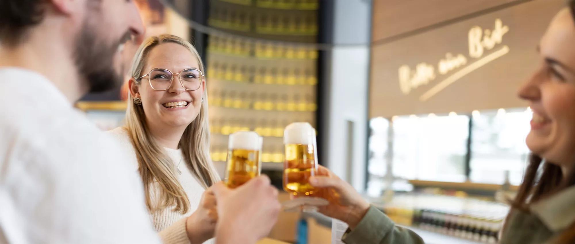 Drei Besucher stoßen in der Bit-Lounge mit frisch gezapftem Bitburger Bier an. Der Hintergrund ist verschwommen. Rechts ist die Leuchtschrift mit dem Markenslogan "Bitte ein Bit" zu erkennen.