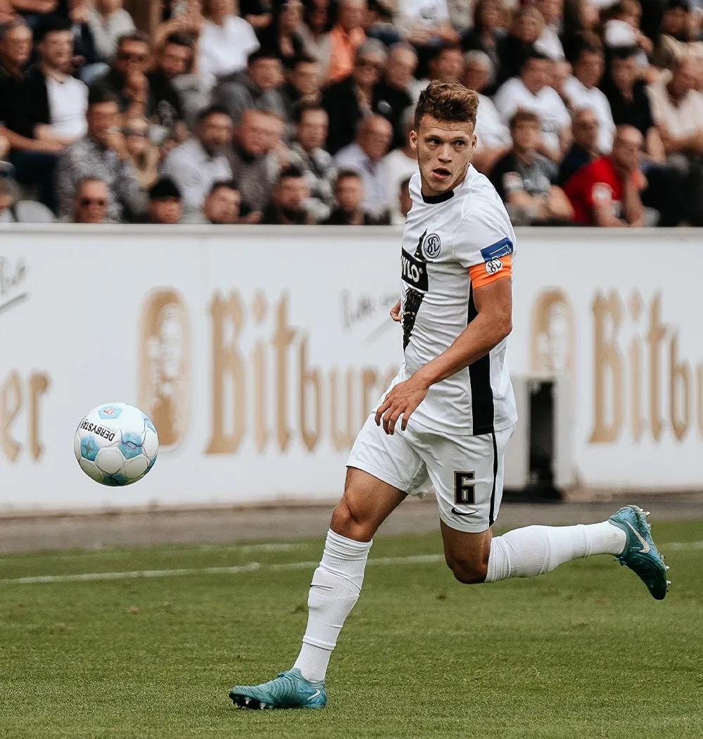 Ein Fußballspieler ist auf dem Spielfeld und in der Bewegung, einen Ball zu schießen. Dabei schaut er in die Schießrichtung.