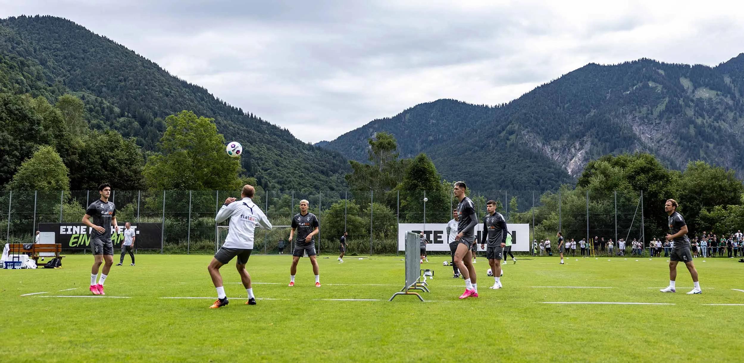 Foto vom Trainingslager Borussia Mönchengladbach. Spieler spielen Fußball auf dem Fußballfeld. Im Hintergrund ist eine bergige Landschaft zu sehen.