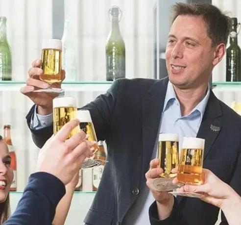 Ein Mann im dunklen Sakko stößt mit mehreren Personen mit frisch gezapftem Bitburger im Glas an. Im Hintergrund stehen dekorative Bierflaschen in einem Regal. Die Szene wirkt fröhlich und gesellig.