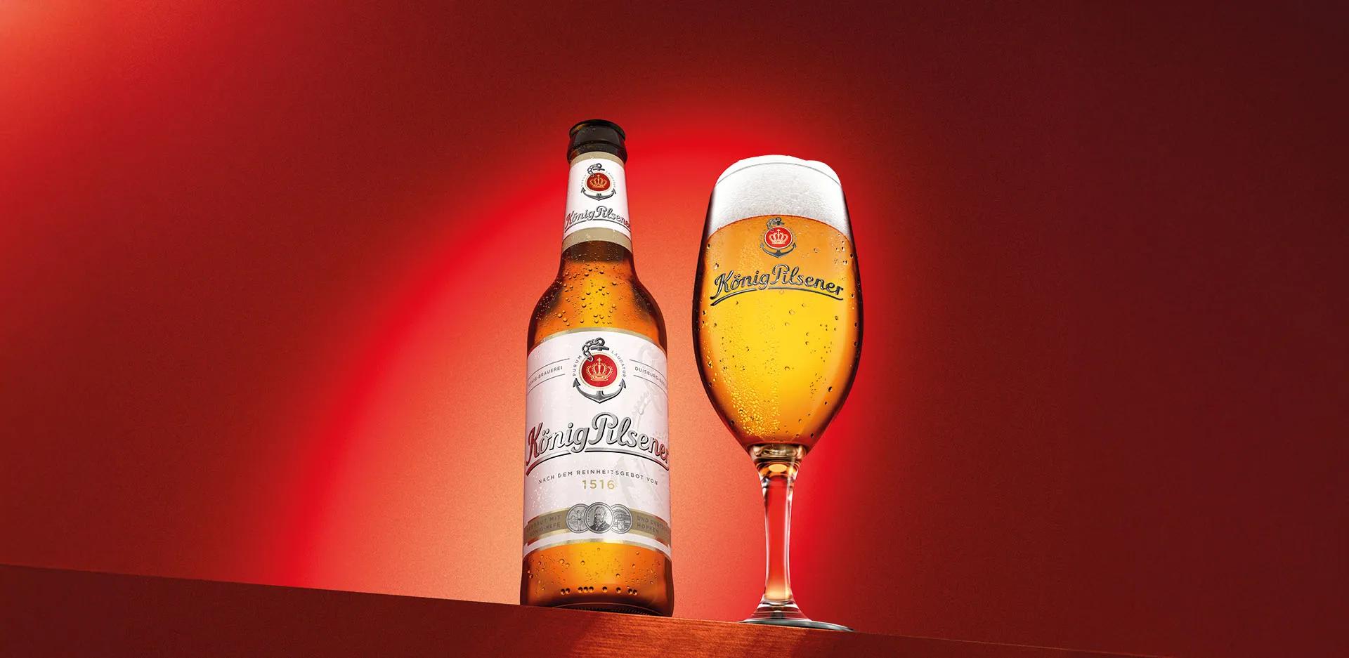 Eine König Pilsener Flasche steht neben einem vollen König Pilsener Glas auf dunkelrotem Hintergrund.