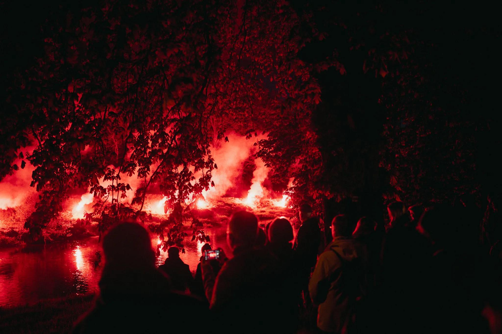 Rötliche Fackeln erleuchten die Nacht, im Vordergrund sieht man eine Menschenmenge, die zuschaut.