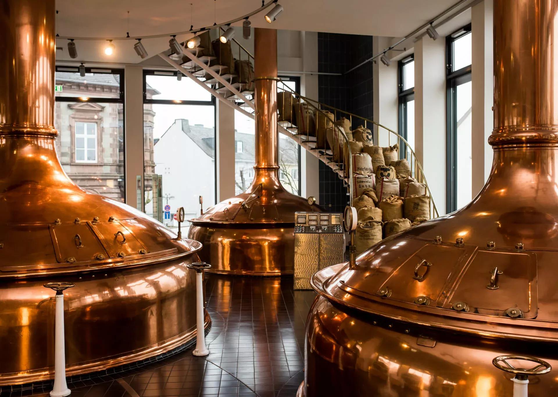 Das historische Sudhaus der Bitburger Brauerei mit glänzenden Kupferkesseln. Rechts führt eine Treppe nach oben, die nichz begehbar ist, da diese mit vollen Jutesäcken dekorativ belagert ist. Im Hintergrund sieht man deckenhohe Fenster.