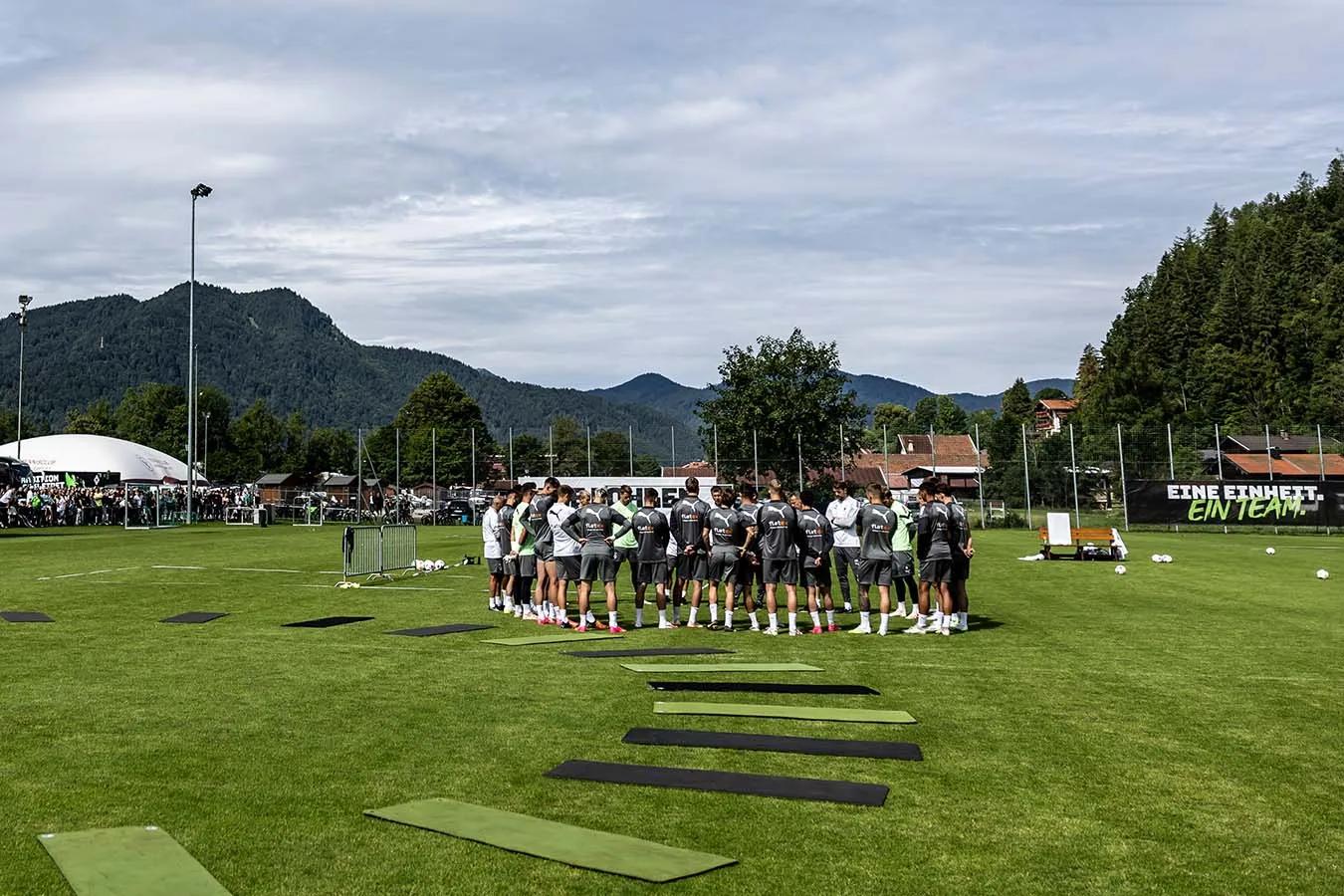 Foto vom Trainingslager Borussia Mönchengladbach. Spieler stehen auf dem Fußballfeld und besprechen sich. Im Hintergrund ist eine bergige Landschaft zu sehen.