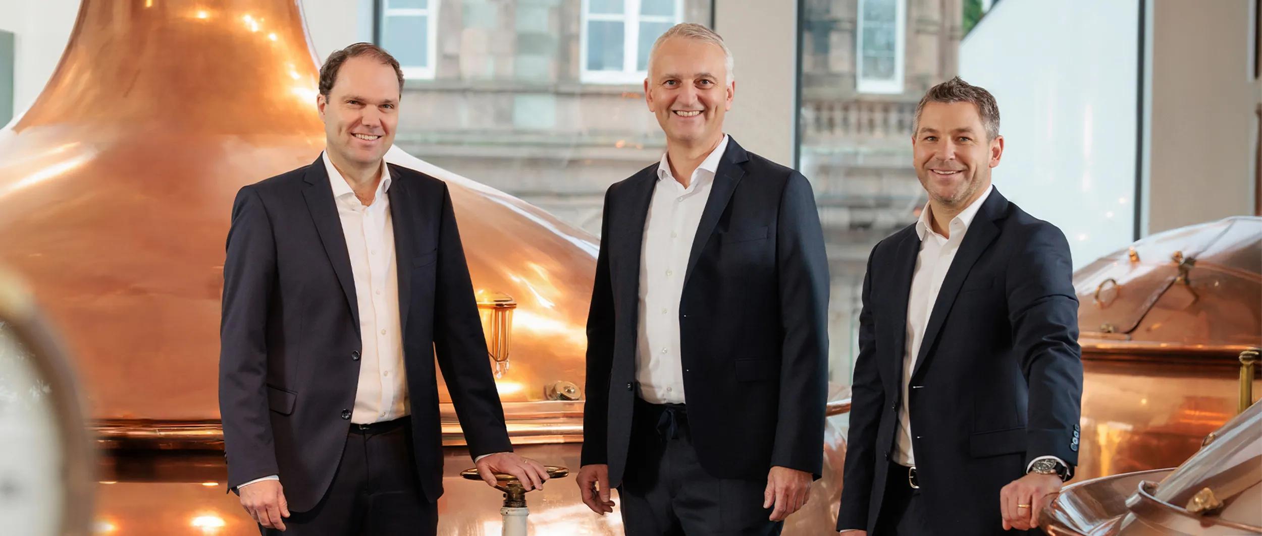 Drei Geschäftsführer in dunklen Anzügen stehen vor großen Kupferkesseln in einer Brauerei und lächeln in die Kamera.