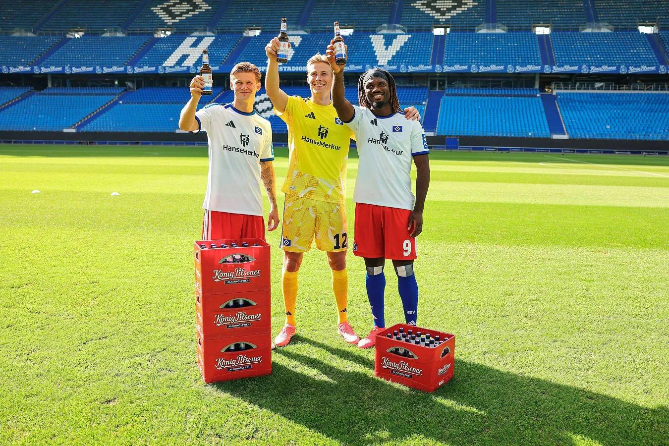 Teamfoto HSV : 3 Fußballer stehen auf dem Rasen mit 4 Kästen König Pilsener und prosten der Kamera mit je einer Flasche Bier in der Hand zu.