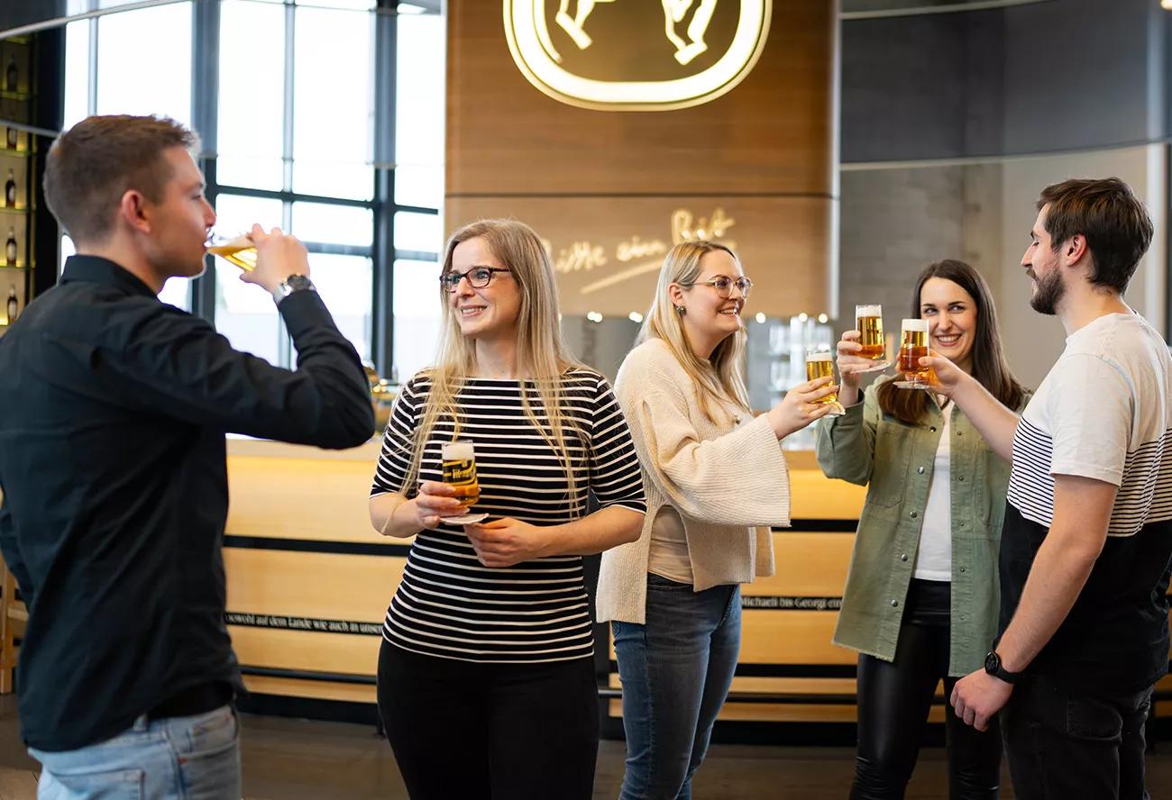 Fünf Besucher der Bitburger Erlebniswelt stoßen in der Bit-Lounge mit frisch gezapftem Bitburger Bier an. Im Hintergrund ist verschwommen die Theke zu sehen. 