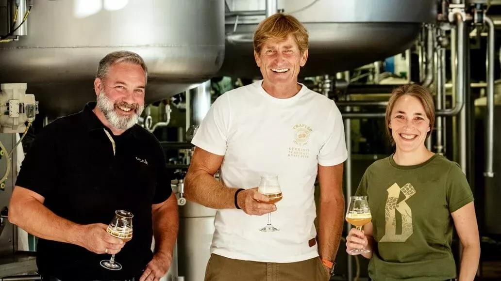 Zwei Männer und eine Frau stehen in einer Bierbrauereianlage und halten ein gefülltes Bierglas in der Hand.