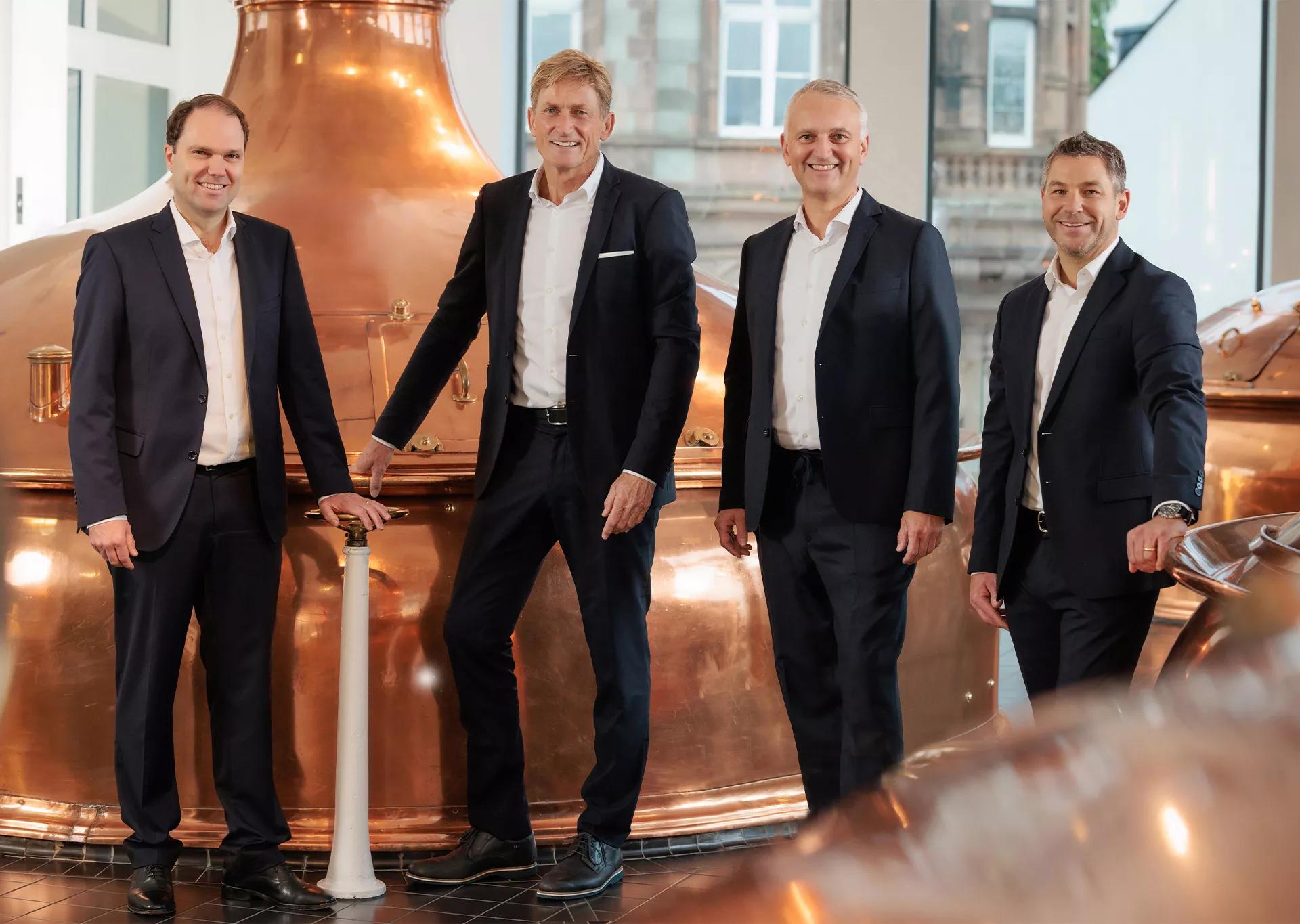Geschäftsführung der Bitburger Braugruppe, vier Männer im Anzug stehen am Kupferkessel
