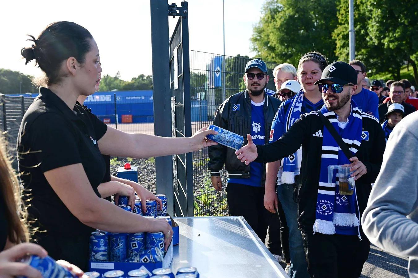 Fans vom HSV stehen in einer Schlange an, eine Person überreicht jeweils eine HSV Fandose.