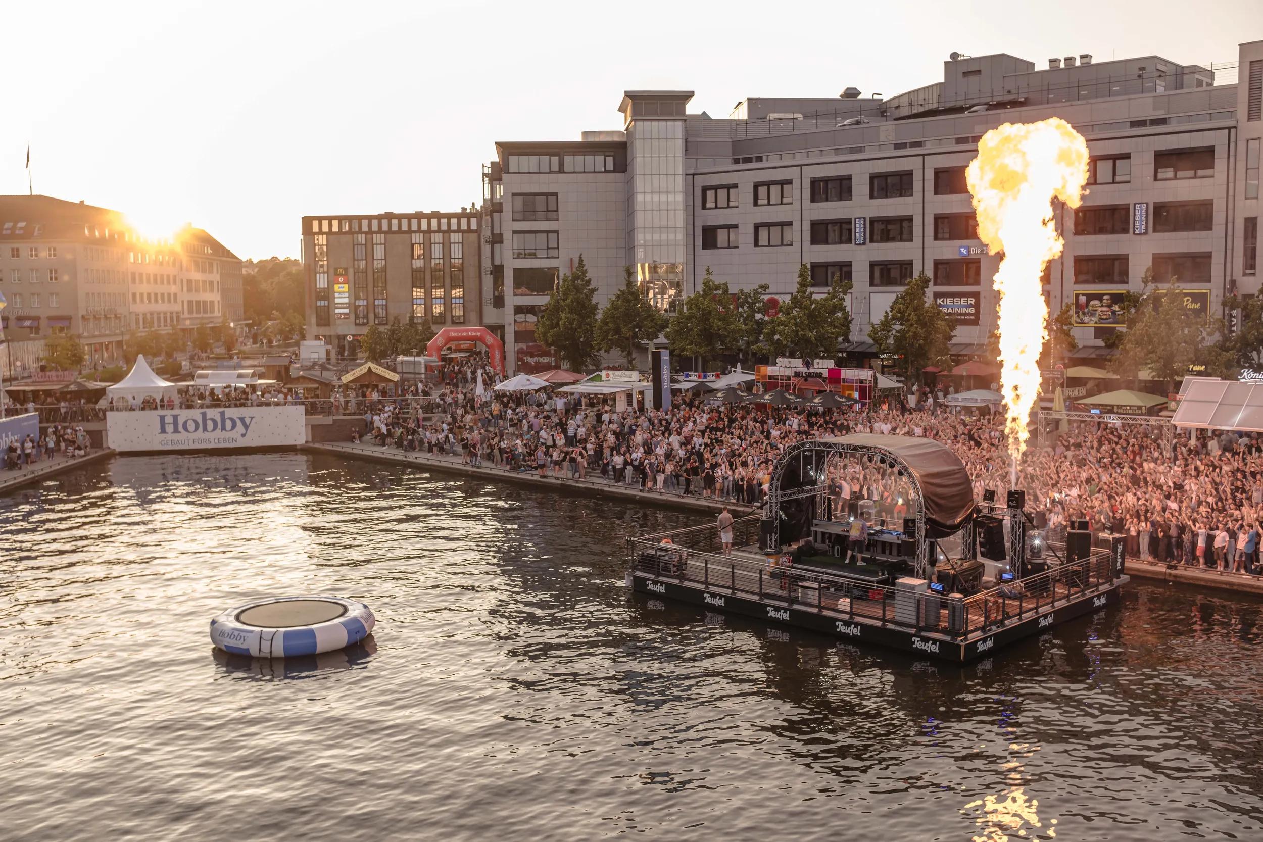 Vogelperspektive auf den Kieler Hafen mit einer feiernden Menschenmenge und einem Feuerspiel, auf dem Wasser vor dem Hafen sieht man ein blau-weißes Luftboot