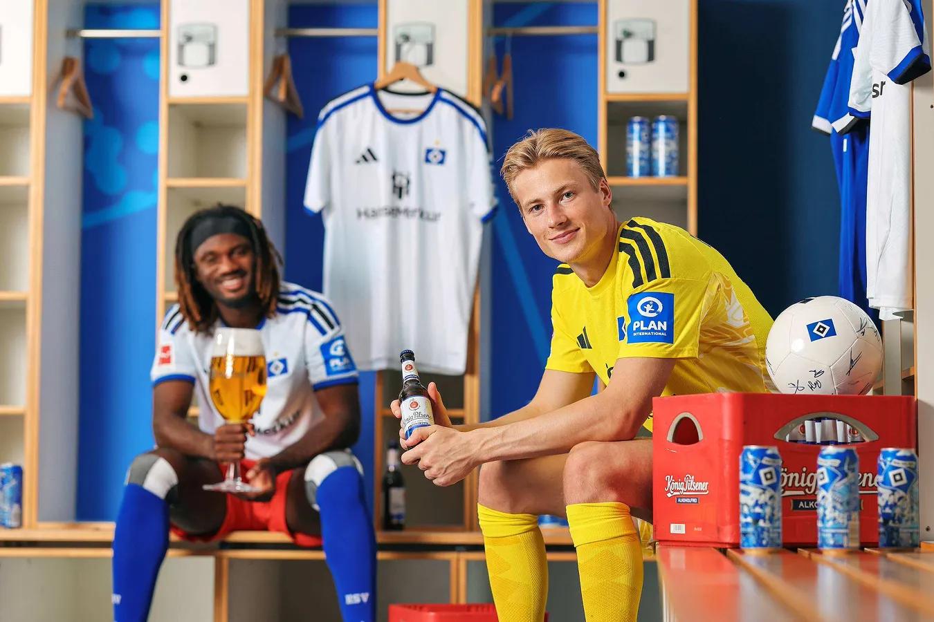 Teamfoto HSV: Zwei Fußballer in der Umkleidekabine sitzend, lächeln in die Kamera und haben ein Bier in der Hand.