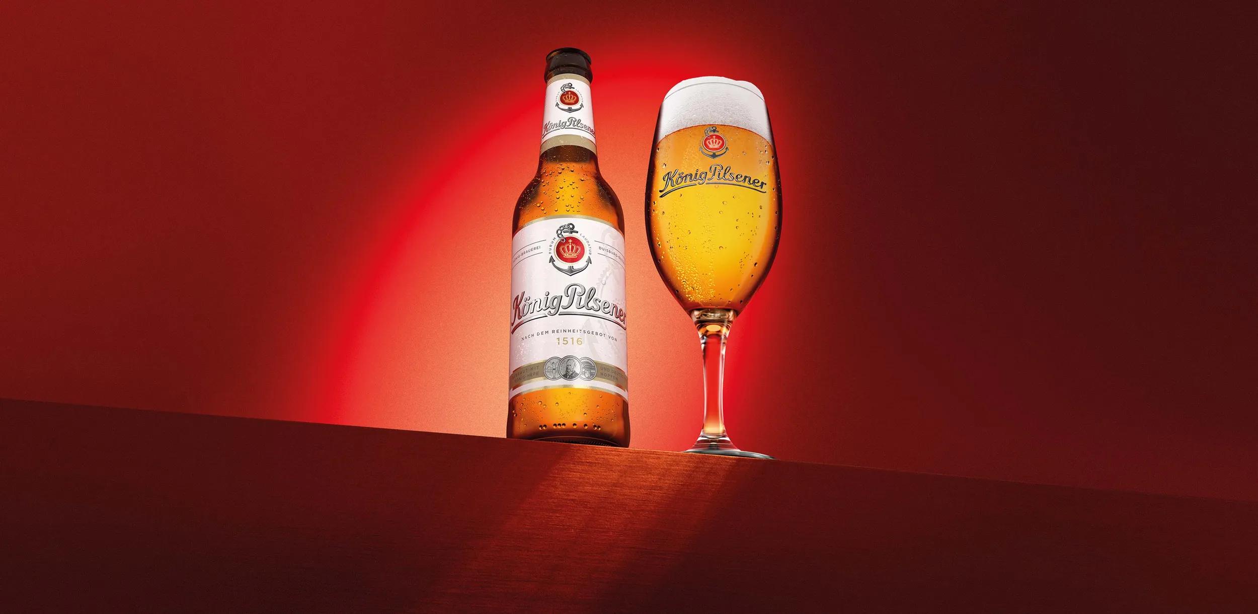 König Pilsener Flasche und gefülltes Glas mit Schaumkrone vor leuchtend rotem Hintergrund.