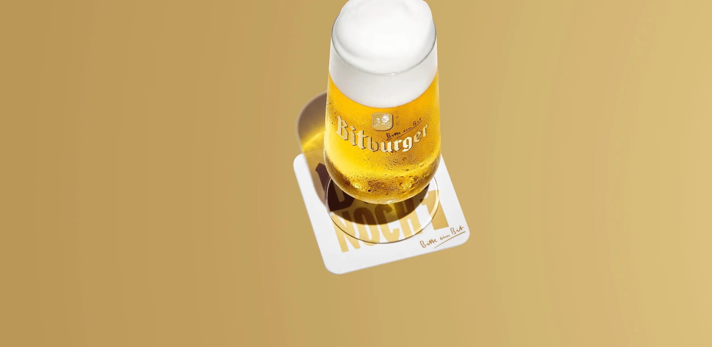 Ein frisch gezapftes Bitburger Pilsener im Glas mit Schaumkrone auf einem Bierdeckel, vor goldenem Hintergrund.