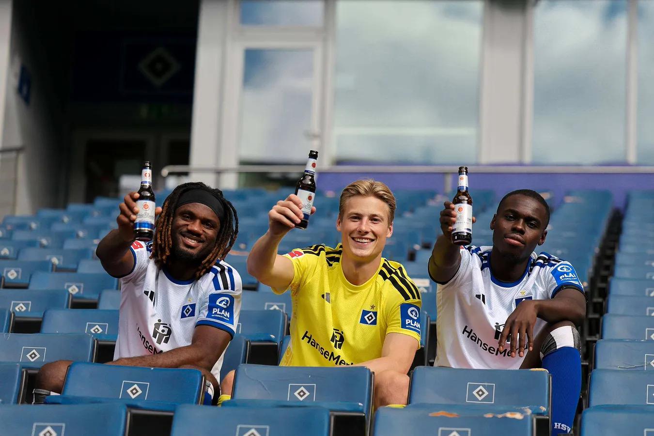 Teamfoto HSV: 3 Fußballer sitzen alleine auf der Tribühne und prosten der Kamera mit je einer Flasche Bier in der Hand zu.