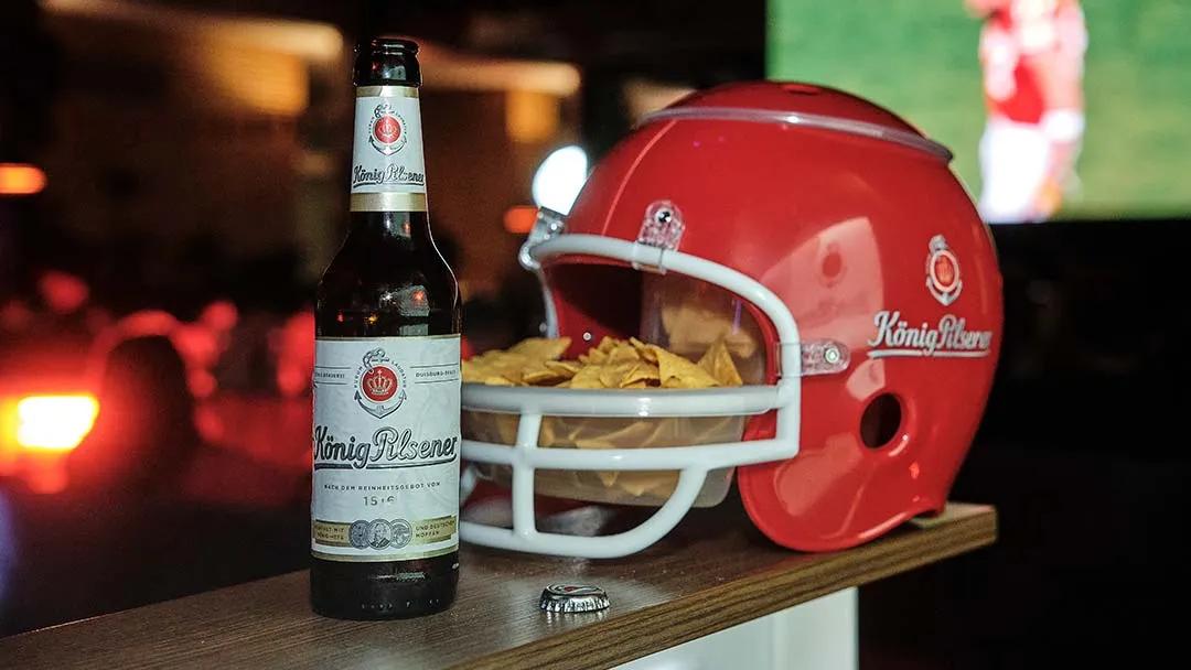 Roter Football Helm und König Pilsener Flasche auf einem Tisch in einer Bar, Im Hintergrund ist ein Fernseher mit einem Football Spiel