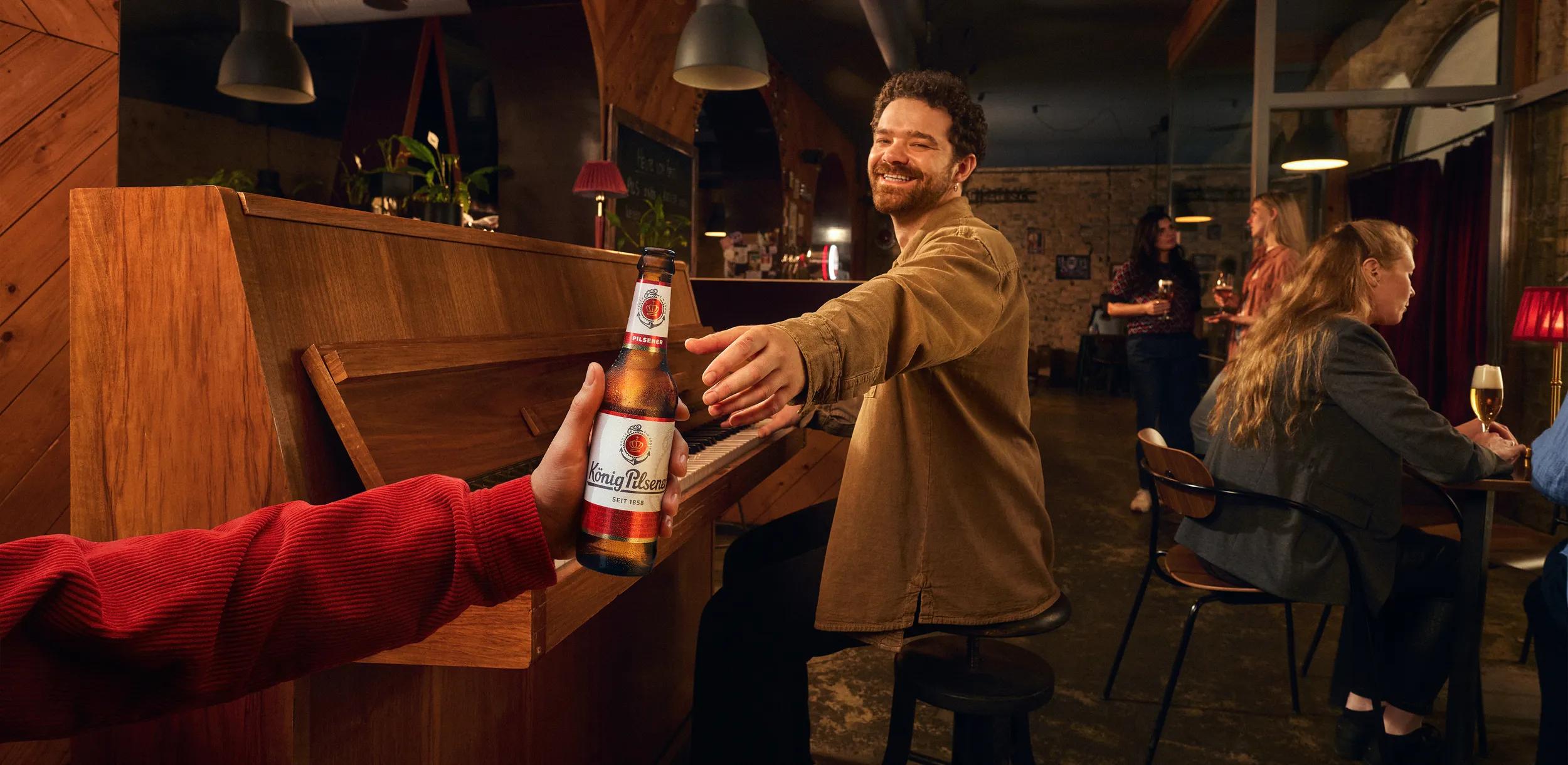 Einer Person an einem Klavier in einer Kneipe wird ein Bier gereicht.