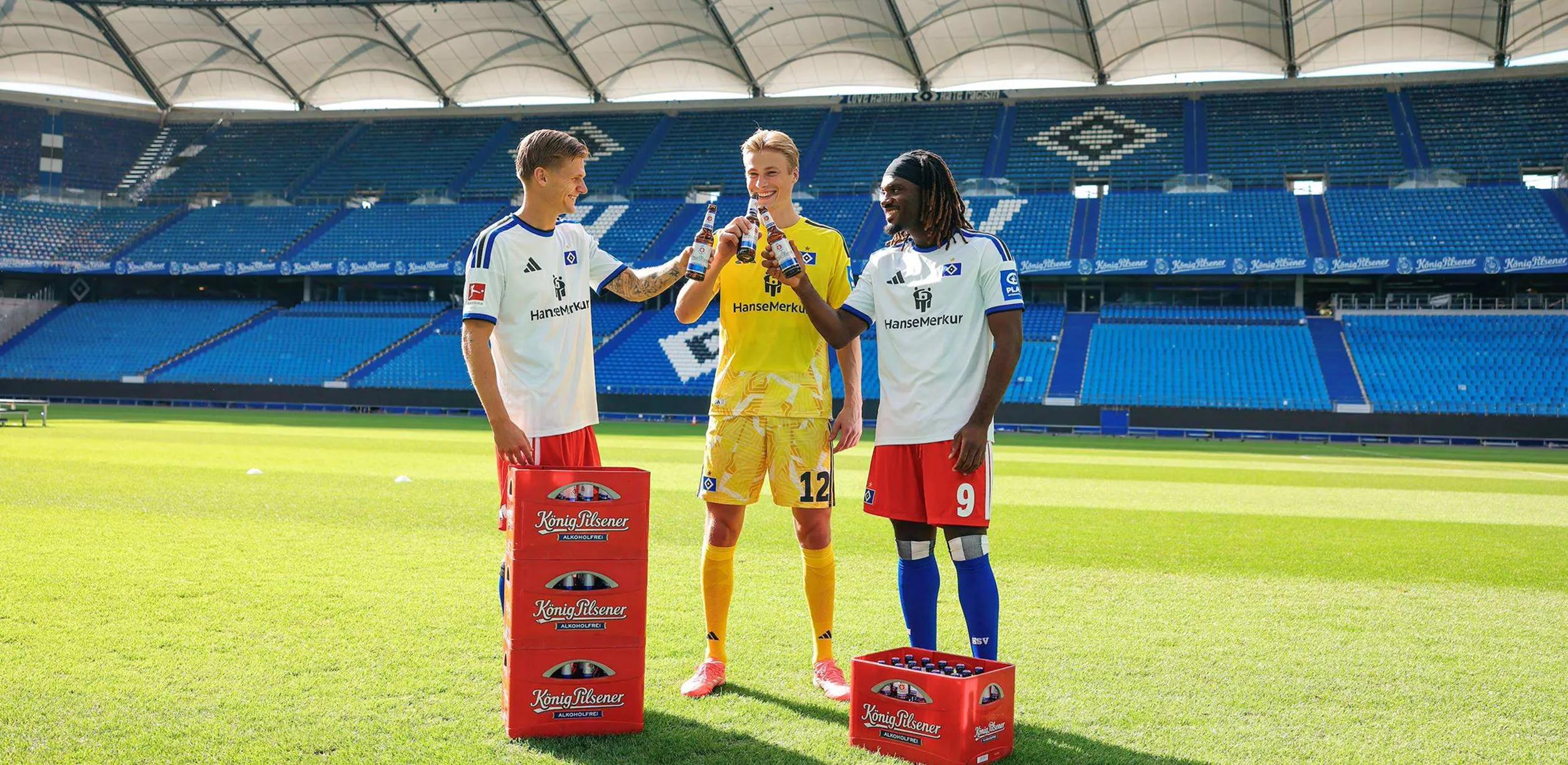 Teamfoto HSV : 3 Fußballer stehen auf dem Rasen mit 4 Kästen König Pilsener und stoßen mit je einer Flasche Bier in der Hand an.