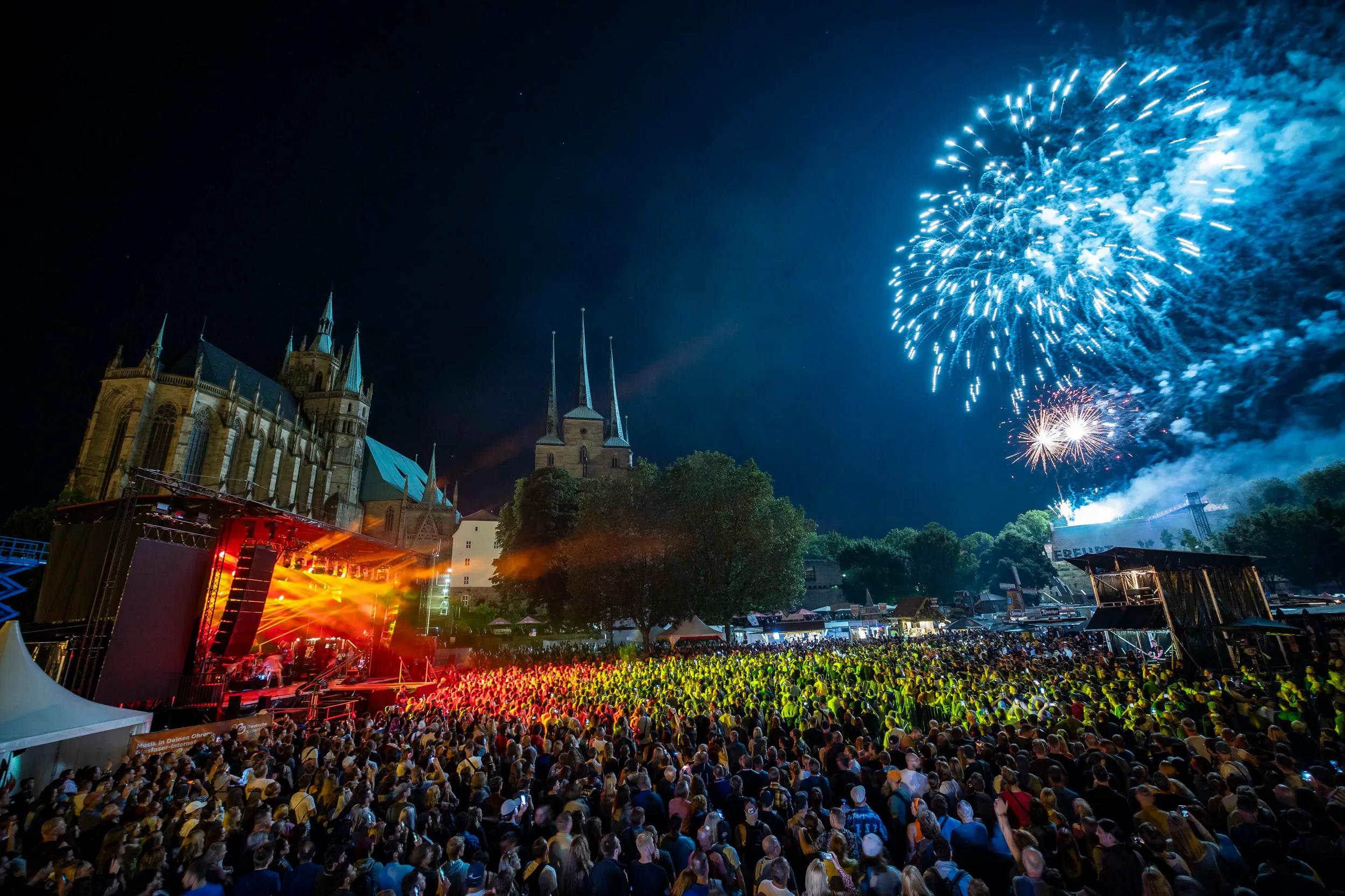 Große Menschenmenge steht vor einer Musikbühne, die bunt beleuchtet wird. Im Hintergrund ist eine große Kirche und Feuerwerk zu sehen.