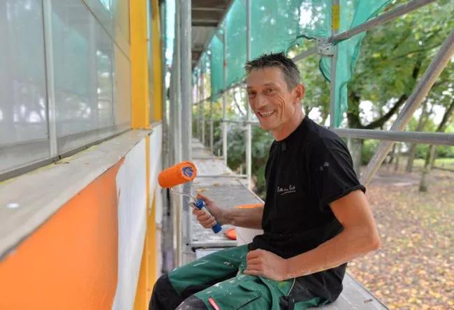 Trier Aktiv im Team Mann sitzt auf Baugerüst und streicht die Außenfassade mit orangener Farbe