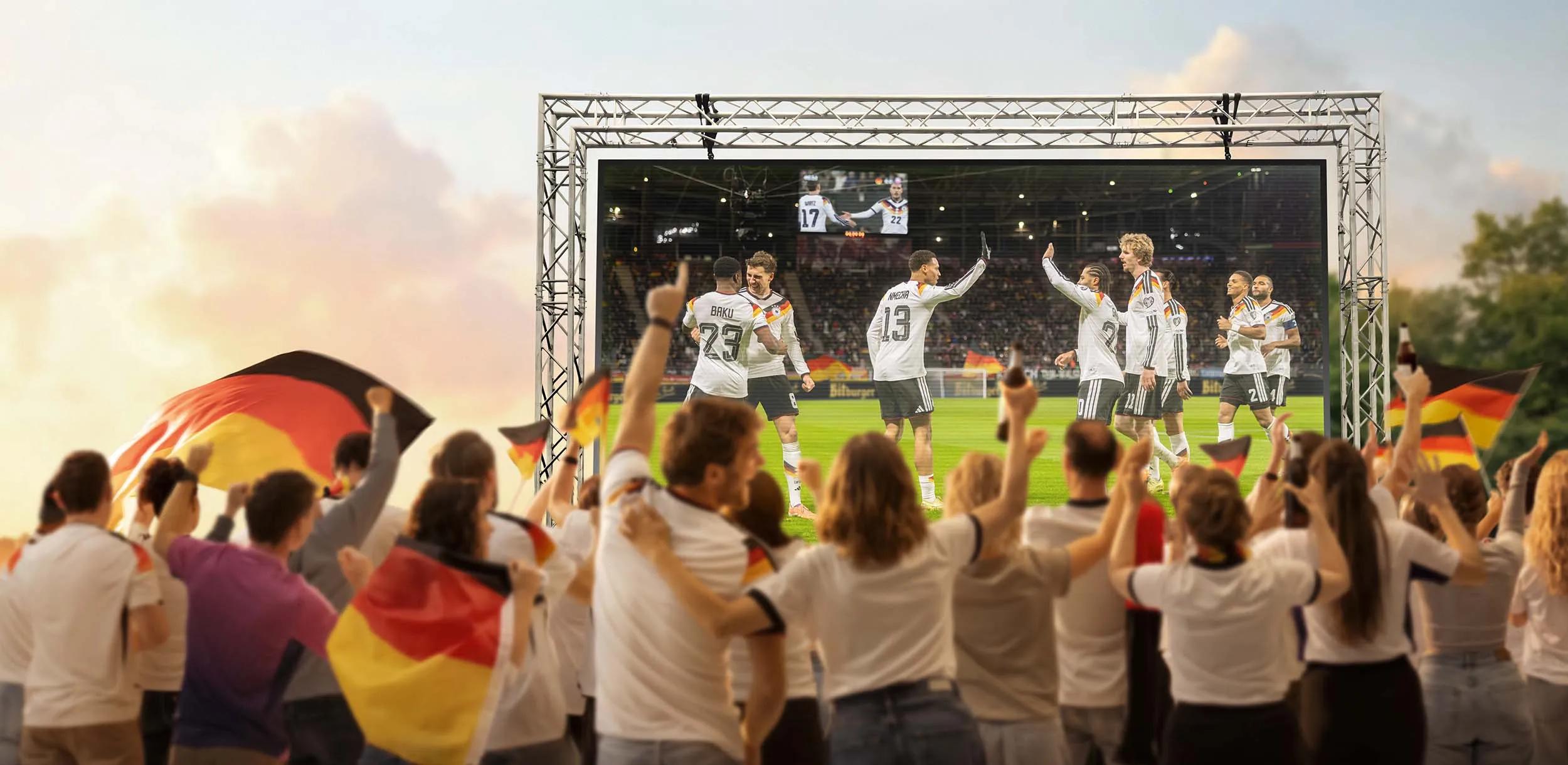 Eine Gruppe von feiernden Fußballfans von hinten, die gerade ein Fußballspiel auf einer Leinwand schauen.