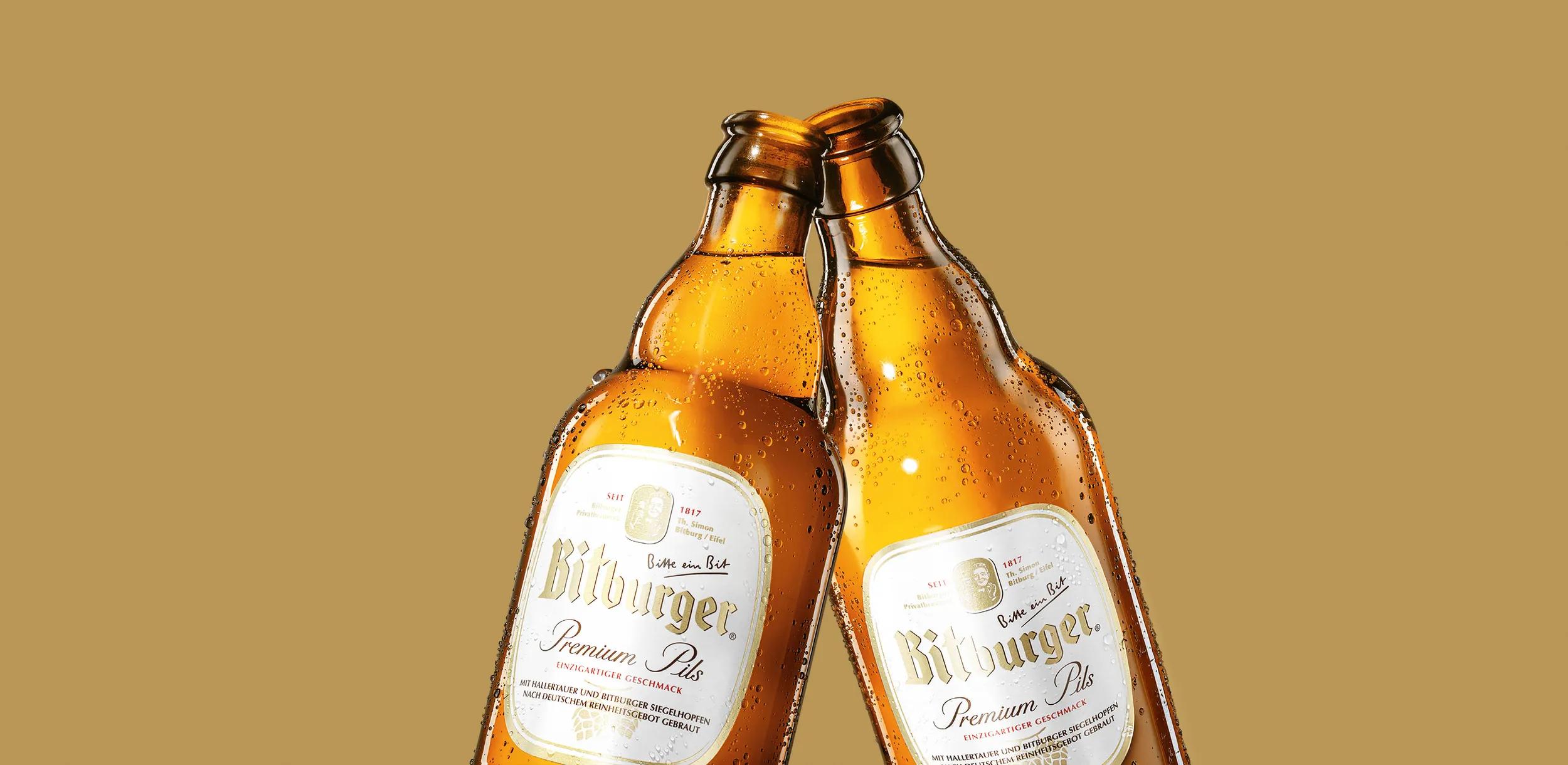 Zwei Bitburger Stubbi-Bierflaschen stoßen an vor goldenem Hintergrund.