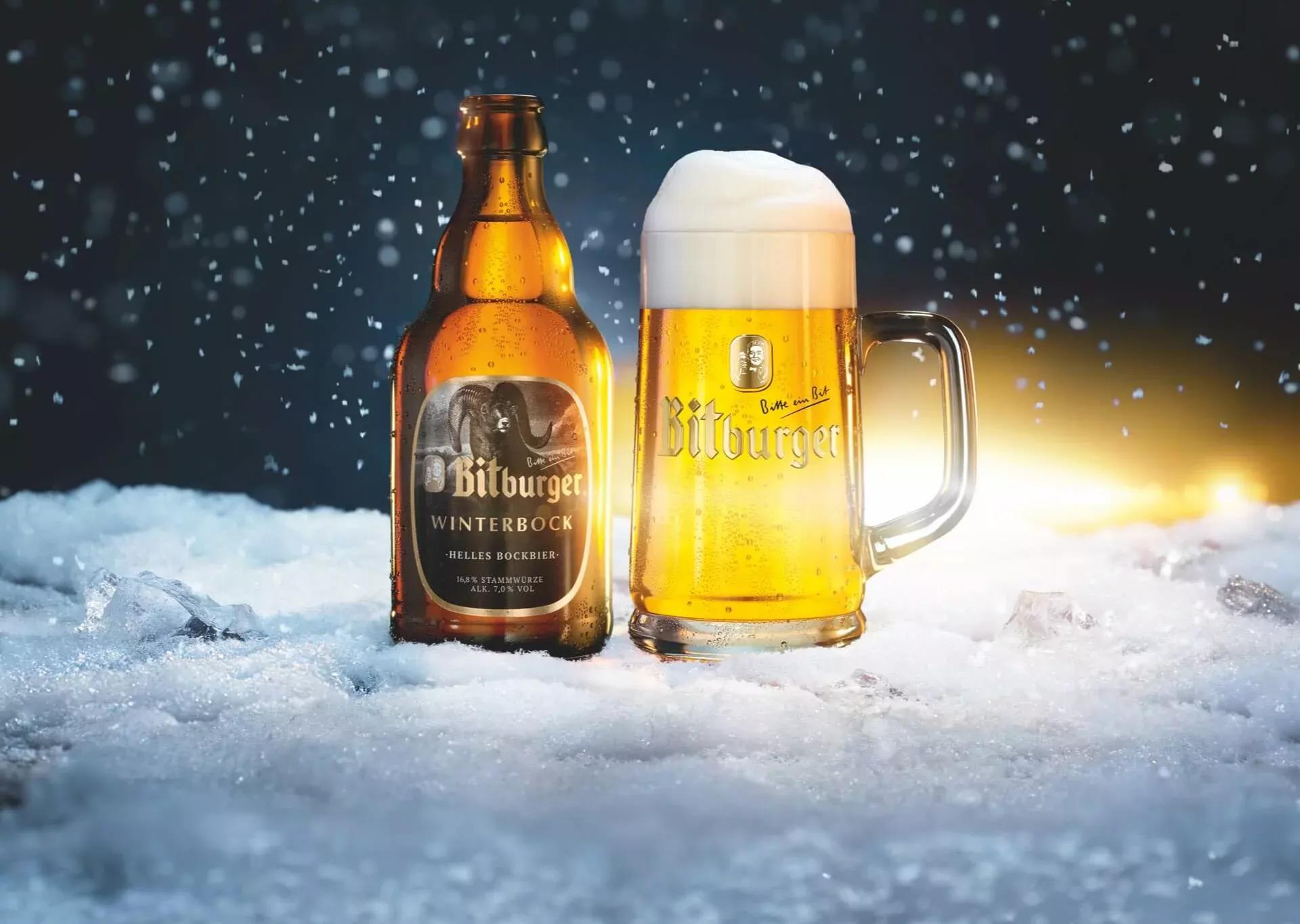 Stubbi Flasche Winterbock mit einem Bierkrug gefüllt mit Bitburger Winterbock auf schneebedecktem Boden, im Hintergrund Nachthimmel und es schneit.
