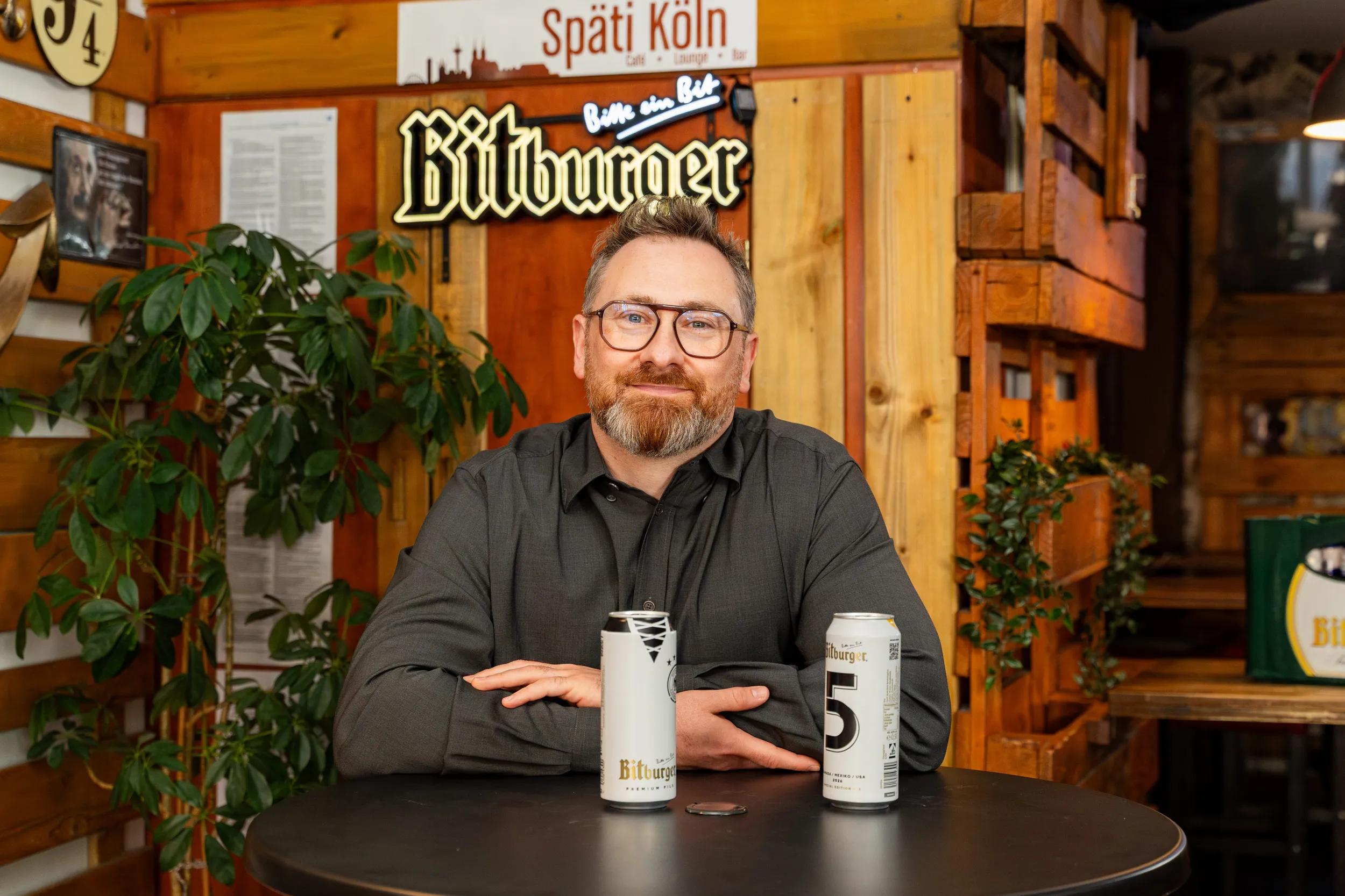 Mann stützt sich auf einen Tisch mit zwei Bitburger Dosen und lächelt in die Kamera.