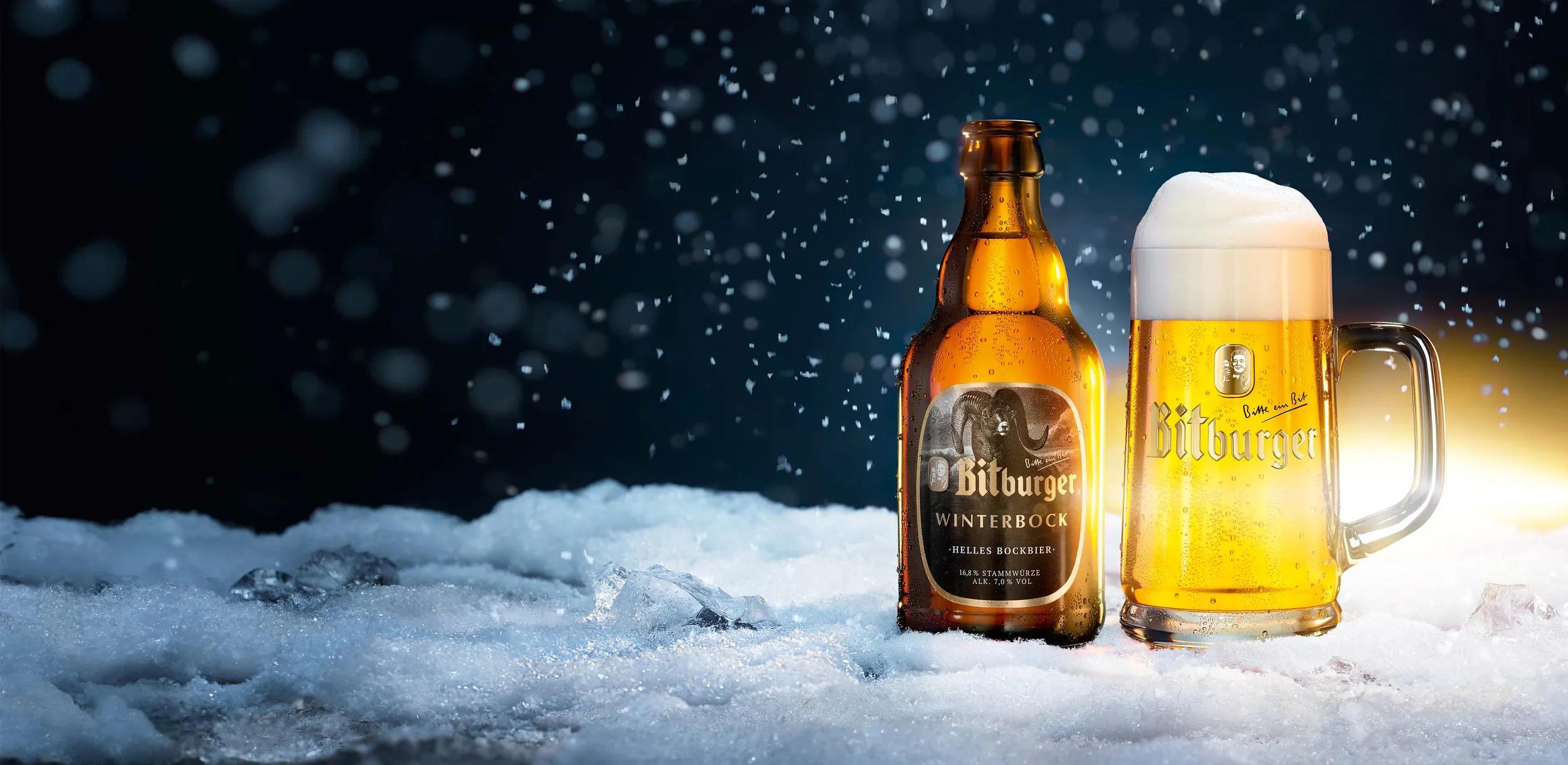 Stubbi Flasche Winterbock mit einem Bierkrug gefüllt mit Bitburger Winterbock auf schneebedecktem Boden, im Hintergrund Nachthimmel und es schneit.